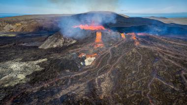 A dramatic aerial view of a volcanic crater with molten lava flows, glowing orange channels on dark, rugged lava, and wisps of smoke rising into a pale blue sky over a barren landscape.