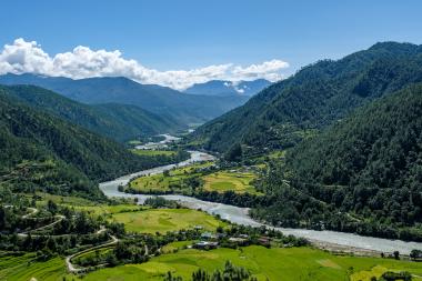 Panoramic landscape view in the Punakha region of Bhutan: a broad, forested mountain valley with a winding river cutting through bright green terraced farmland and scattered village houses beneath a clear blue sky with a few white clouds.