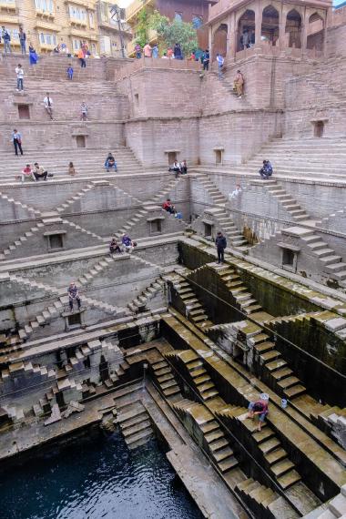 Wide, top-down view into Toorji Ka Jhalra stepwell in Jodhpur, Rajasthan, showing geometric stone staircases and terraces descending to a dark pool of water. Several visitors sit on the steps, and one man crouches near the lower steps with a bucket, washing beside the water. Photo taken in the morning on 21 Dec 2023 near 26.297325, 73.022983.