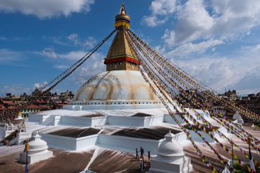 Large white stupa with a golden spire and colorful prayer flags radiating outward from its top, set against a blue sky with scattered clouds; surrounding city buildings and a few visitors at the base.