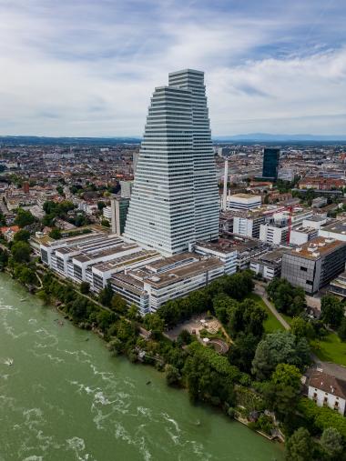 A drone-shot of a tall, tiered white office building near the Rhine river in Basel, Switzerland. The surrounding cityscape stretches into the distance under a partly cloudy blue sky, with green trees along the riverbank and a park area at the base of the building.