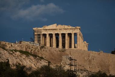 A view of the Parthenon on the Acropolis in Athens, with ongoing restoration scaffolding around the columns, a rocky hillside below, and a deep blue sky above.