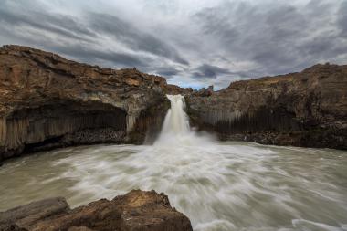 Long-exposure photograph of a waterfall dropping between dark basalt cliffs with columnar rock formations, churning milky water at the base, and a cloudy sky overhead in Iceland.