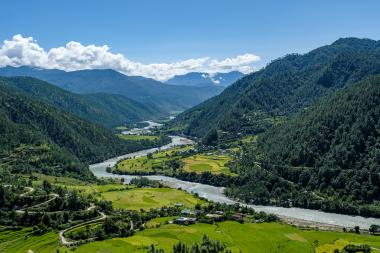 Panoramic landscape view in the Punakha region of Bhutan: a broad, forested mountain valley with a winding river cutting through bright green terraced farmland and scattered village houses beneath a clear blue sky with a few white clouds.