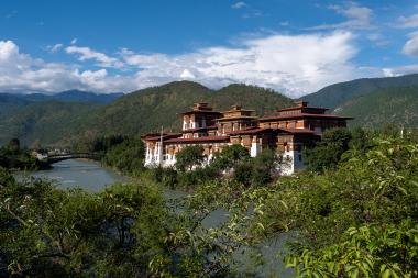 Wide landscape view of Punakha Dzong, a large white-and-red Bhutanese fortress monastery with tiered golden roofs, perched beside the Mo Chhu River; green shrubs fill the foreground, a bridge crosses the river to the left, and forested mountains rise under a blue sky with scattered clouds in Punakha, Bhutan.