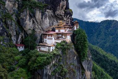 Wide landscape view of Paro Taktsang (Tiger’s Nest) monastery in Bhutan: white-and-red temple buildings with golden roofs built into a vertical rock face, surrounded by dark green conifer forest and steep mountains, with heavy gray clouds overhead at dusk.