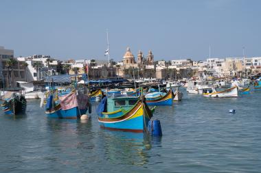 A wide view of Marsaxlokk harbour showing numerous colorful traditional Maltese fishing boats (luzzus) floating on calm water in the foreground. Behind them is the low-rise coastal village with palm trees and a prominent ochre church dome and twin bell towers on the skyline. Small motorboats and harbour structures line the waterfront under a clear blue sky.