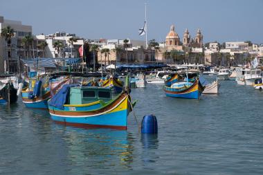 Wide-angle view of a calm harbour filled with colorful traditional Maltese fishing boats (luzzus) painted blue, yellow and green, tied to buoys in the foreground. Behind the boats are waterfront cafés and low stone buildings lined with palm trees, and a large domed church and twin bell towers rising above the town. Clear blue sky and gentle ripples on the water convey a sunny, peaceful day at Marsaxlokk harbour, Malta.