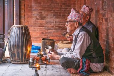 An elderly man with a patterned cap, glasses, and a gray shirt sits cross-legged on a woven mat against a red brick wall. He holds a small metal horn or bell while another man behind him plays a similar instrument. A large wooden ceremonial drum stands to the left, and several small oil lamps and metal vessels are placed on the ground, creating a warm, amber glow.