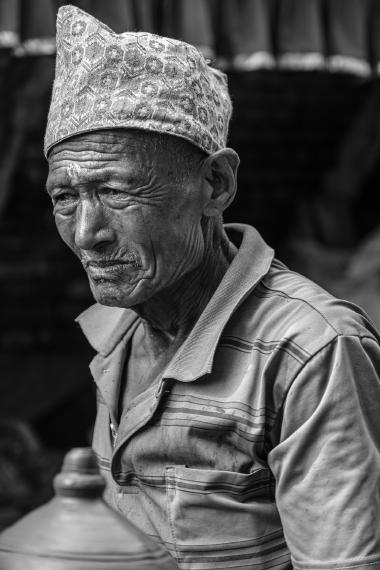 Close-up black-and-white portrait of an older man wearing a patterned traditional cap and a striped shirt, looking slightly downward with a thoughtful, weathered face at a street market.