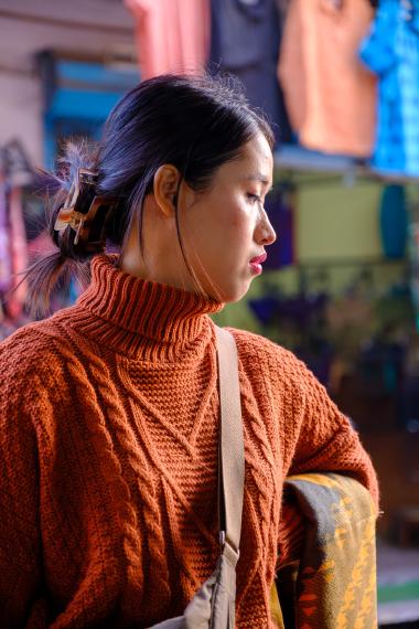 A side-profile portrait of a woman wearing an orange cable-knit turtleneck sweater and a crossbody strap, standing indoors at a market stall with blurred, brightly colored hanging fabrics in the background (Pushkar, Rajasthan, India).