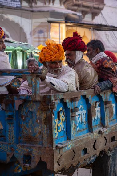 Evening street scene in Pushkar, Rajasthan: several adult men wearing traditional colorful turbans sit and stand in the open bed of an old blue, rusted, hand-painted truck. One man in an orange turban leans forward holding the side rail, while others in red and patterned shawls look to the side. A brightly lit building facade and market-style tarps are visible behind them.