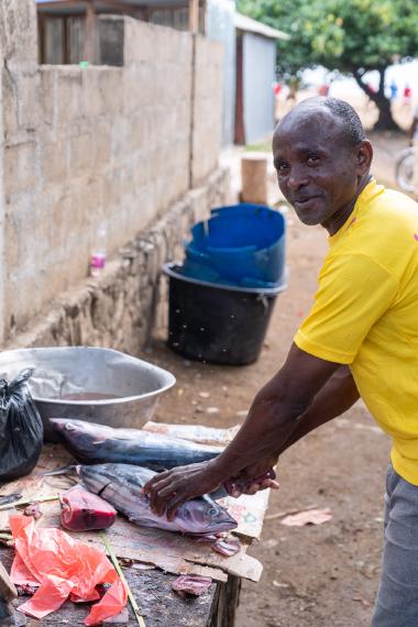 Outdoor roadside fish market scene on Mohéli, Comoros. A man in a bright yellow shirt looks toward the camera while cleaning large tuna on a worn wooden table covered with newspaper; fish pieces, a metal basin, plastic bags, and blue buckets sit nearby along a concrete wall under daylight.